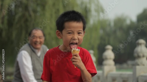 MS Portrait of young boy eating ice cream cone in park, grandfather looking on in background / China