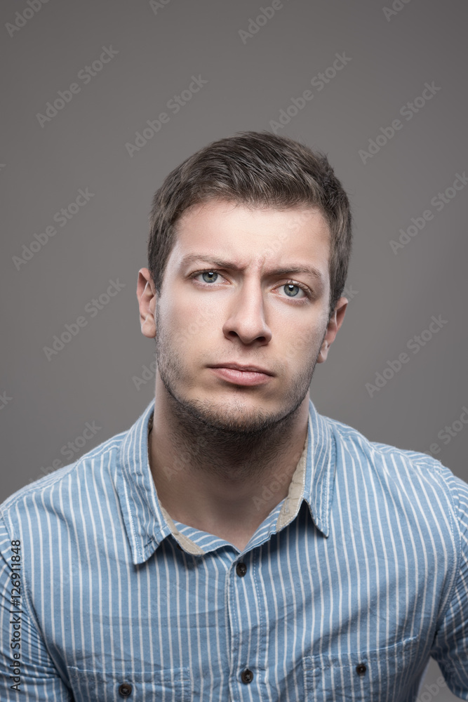 Moody portrait of distrustful young executive man in blue shirt looking at camera over gray background Moody portrait of distrustful young executive man in blue shirt looking at camera over gray background