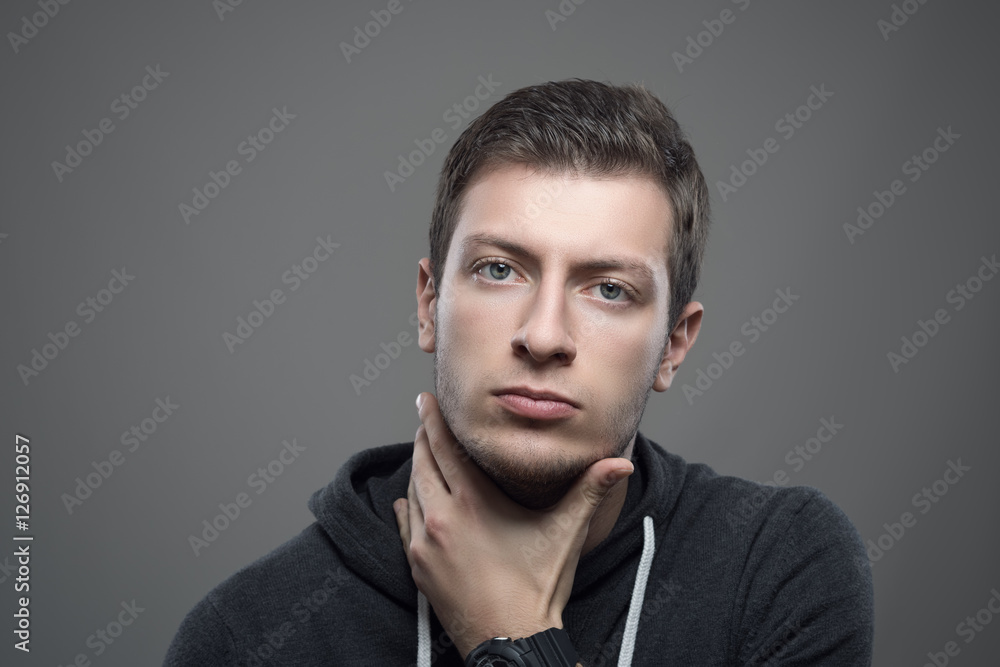 Fototapeta premium Moody portrait of serious young unshaven man with hand under chin looking at camera