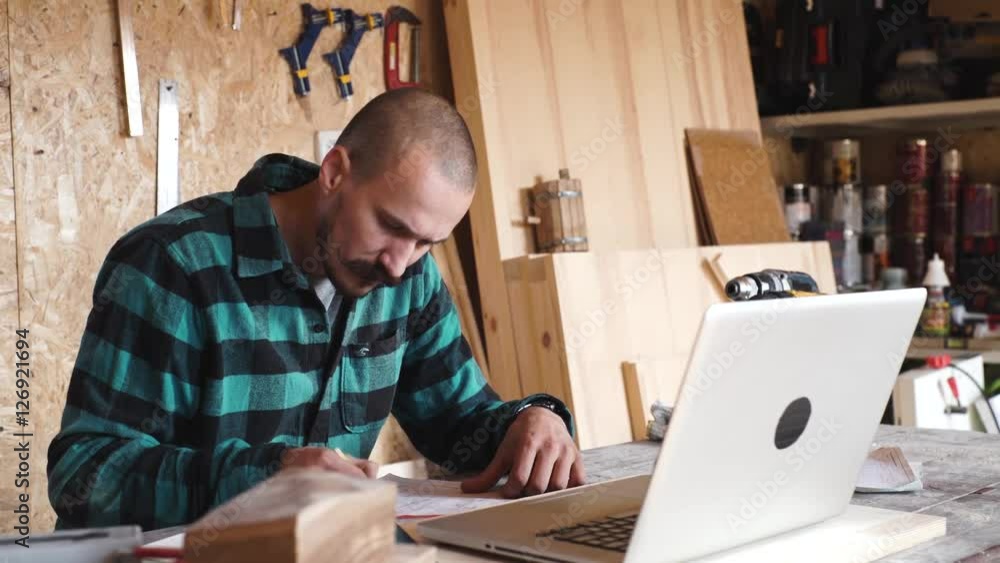 Bold with moustache young Carpenter Craftsman do drawing with laptop in his wooden workshop Bold with moustache young Carpenter Craftsman do drawing with laptop in his wooden workshop