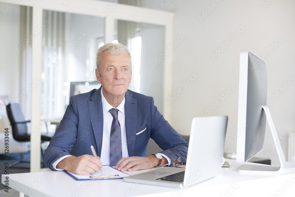 Senior professional businessman at work. Shot of a senior financial manager writing notes while looking at computer at office. Senior professional businessman at work. Shot of a senior financial manager writing notes while looking at computer at office.