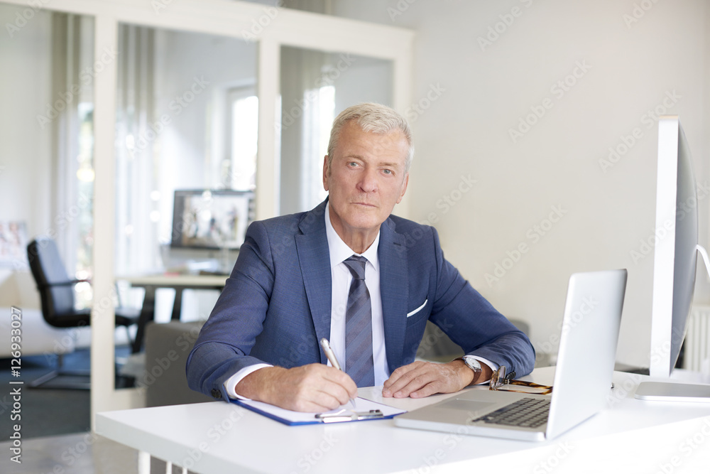 Senior professional businessman at work. Shot of a senior financial manager writing notes while looking at computer at office. Senior professional businessman at work. Shot of a senior financial manager writing notes while looking at computer at office.
