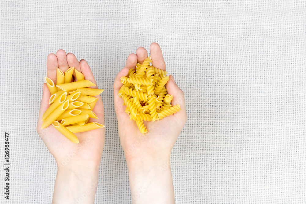 Obraz premium Handful of uncooked pasta flat lay. Female hands holding heap of raw fussilli and penne noodles on white canvas background