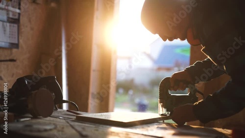 young bold carpenter with moustache. circular saw cutting piece of wood. sun flare on background
