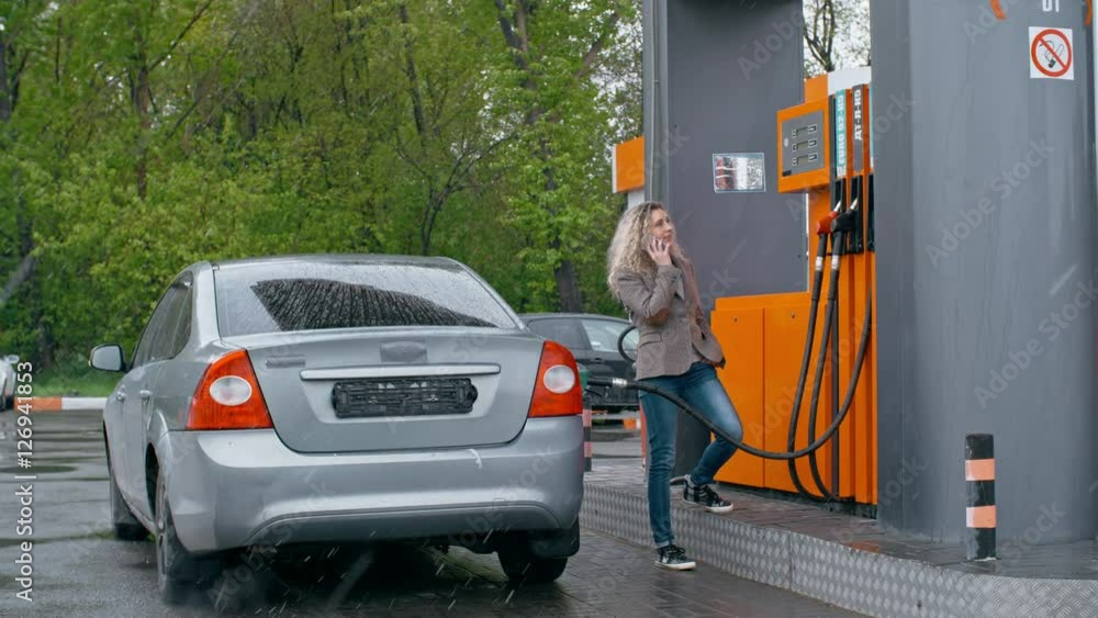 Smiling woman standing at gas station and speaking on cell phone while refilling car with petrol Smiling woman standing at gas station and speaking on cell phone while refilling car with petrol