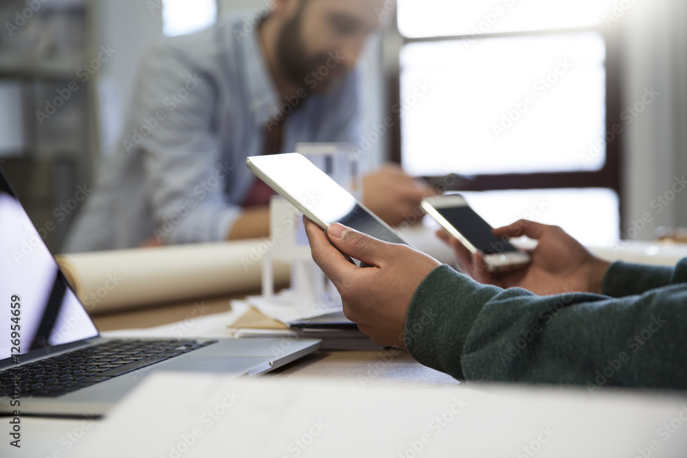 Man with portable devices at desk with colleague in background