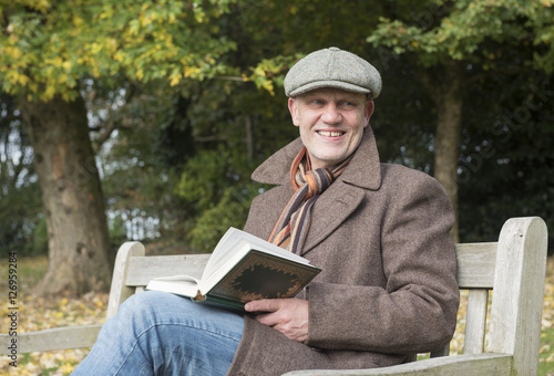 Portrait image of a happy mature man outdoors reading a book on a park bench. 