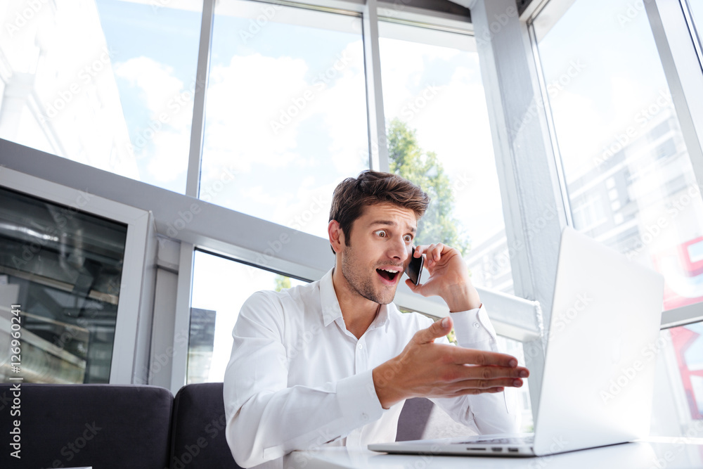 Amazed young businessman using laptop and talking on mobile phone Amazed young businessman using laptop and talking on mobile phone