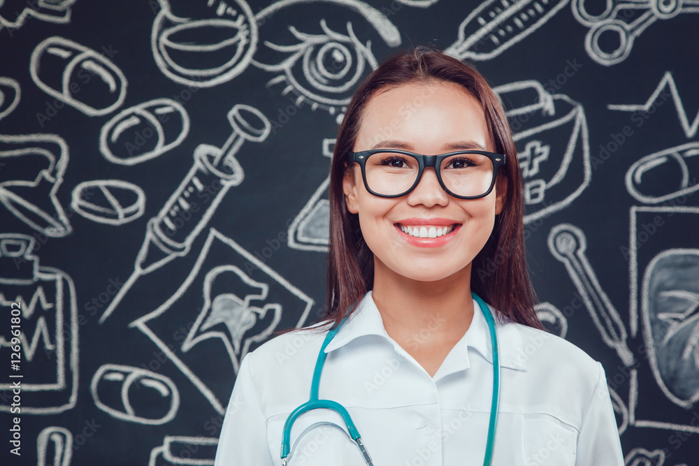 Smiling young woman doctor in glasses and white coat standing on dark background with pattern Smiling young woman doctor in glasses and white coat standing on dark background with pattern