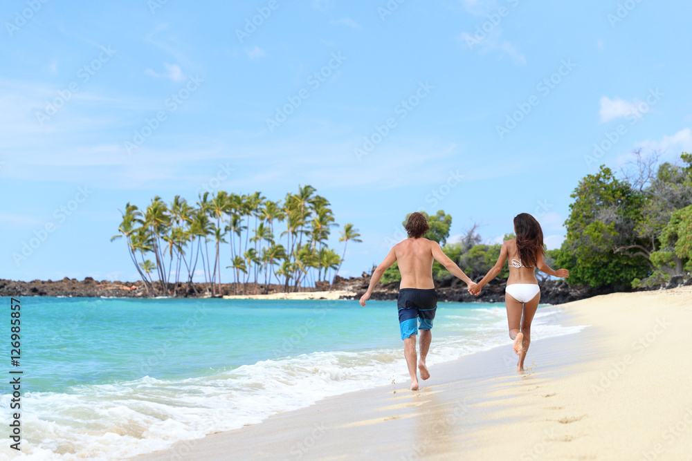 Happy couple from behind holding hands running having fun on tropical beach vacation. Hero view of people walking together in the splashing waves happy on summer holidays.