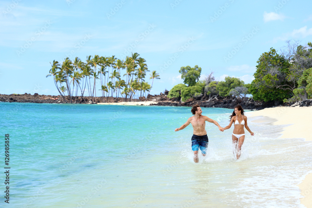 Happy couple laughing together holding hands running having fun splashing water in the ocean waves. Young beautiful fit slim people enjoying their happy lifestyle in paradise destination beach.