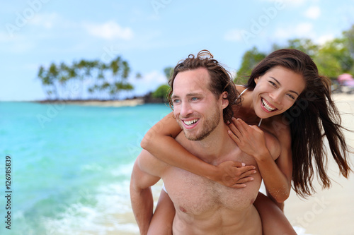 Attractive happy couple laughing having fun piggybacking on tropical beach. Caucasian man carrying Asian girlfriend having fun laughing on travel vacation. Healthy happy interracial people.