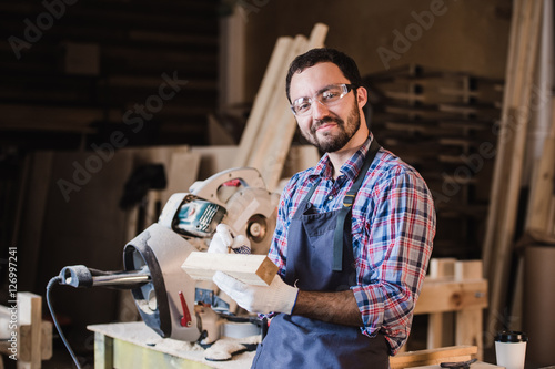 Handsome carpenter in protective glasses is looking at camera and smiling while standing near his wooden object in the workshop
