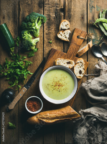 Homemade pea, broccoli, zucchini cream soup in white bowl with fresh baguette on wooden board over rustic background, top view, vertical composition photo