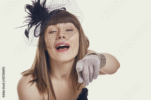 Fényképezés Portrait of an amused beautiful young woman wearing a black vintage style light feather hat, net veil and gloves while pointing finger and laughing at you against bright background