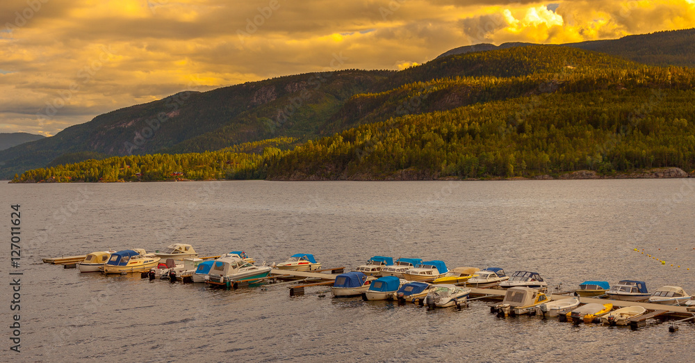 Fototapeta premium Motor speed fishing boats tied with ropes to lake pier in Norway during sunset, Mountains fishing lake with wooden jetty for motorboats, tied to pier. Amazing north mountains lake sunset landscape