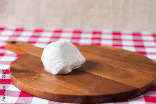 Single water buffalo mozzarella mozzarella di buffala cheese ball on wooden cheese chopping board on red and white checkered tablecloth with brown burlap background with copy space above