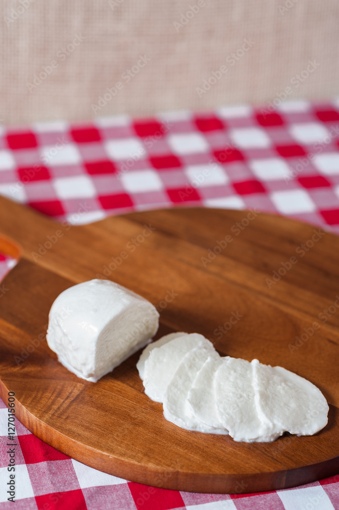Water buffalo mozzarella mozzarella di buffala cheese in ball and sliced on wooden cheese chopping board on red and white checkered tablecloth with brown burlap background with copy space above Water buffalo mozzarella mozzarella di buffala cheese in ball and sliced on wooden cheese chopping board on red and white checkered tablecloth with brown burlap background with copy space above