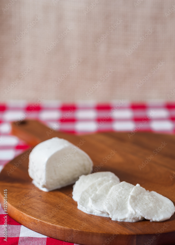 Water buffalo mozzarella mozzarella di buffala cheese in ball and sliced on wooden cheese chopping board on red and white checkered tablecloth with brown burlap background with copy space above Water buffalo mozzarella mozzarella di buffala cheese in ball and sliced on wooden cheese chopping board on red and white checkered tablecloth with brown burlap background with copy space above