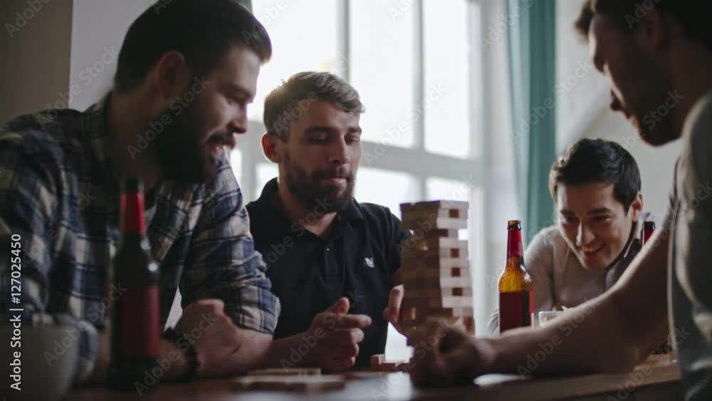 Man carefully taking jenga wooden block from bottom of tower while playing with friends at party Man carefully taking jenga wooden block from bottom of tower while playing with friends at party