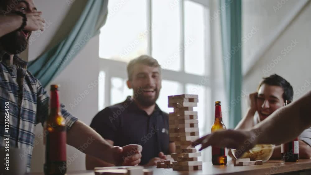 Surprised friends cheering as man taking wooden block from jenga tower without breaking it