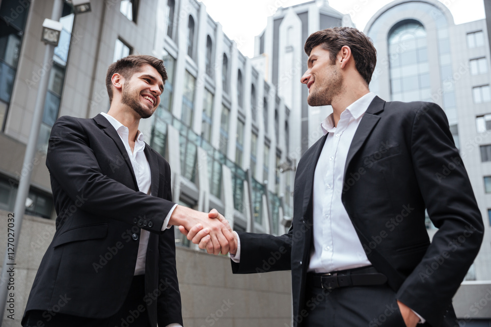 Two happy businessmen standing and shaking hands near business center