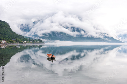 Morning by the fjords, clouds on the mountains with reflection on the water and small boat, Norway