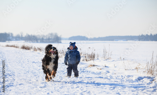 Baby boy and bernese mountain dog running on road at cold winter day 