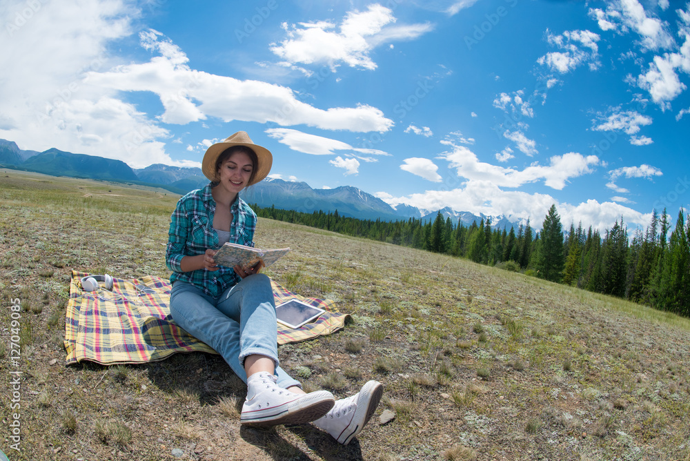 Young woman reading book and sitting in a field. Mountains background.