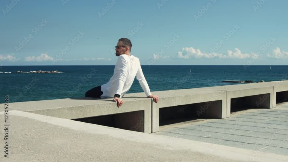 Handsome athletic bearded guy in white longsleeve wearing sunglasses is sitting relaxely on bank next to the sea and looking around in sunny day, camera rotates slowly, seagulls flying on background Handsome athletic bearded guy in white longsleeve wearing sunglasses is sitting relaxely on bank next to the sea and looking around in sunny day, camera rotates slowly, seagulls flying on background