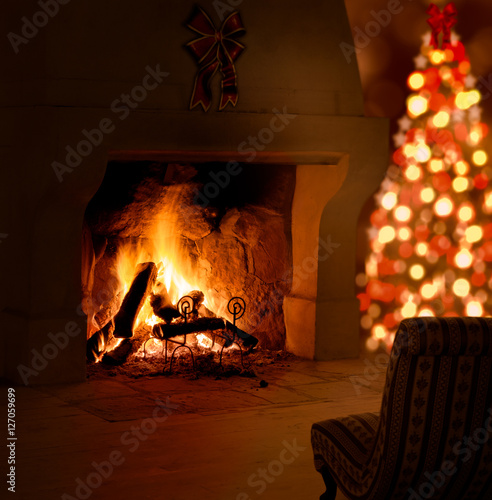 Christmas tree near fireplace. Background with tree, decorations, old armchair, blurry lights and fire burning in a fireplace in a cosy, warm atmosphere in Christmas eve.