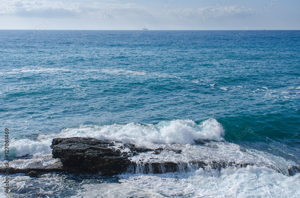 Fototapeta premium Waves hitting a rock near the shore of Nervi, Genoa, in Italy with a boat visible in the distance on the horizon.