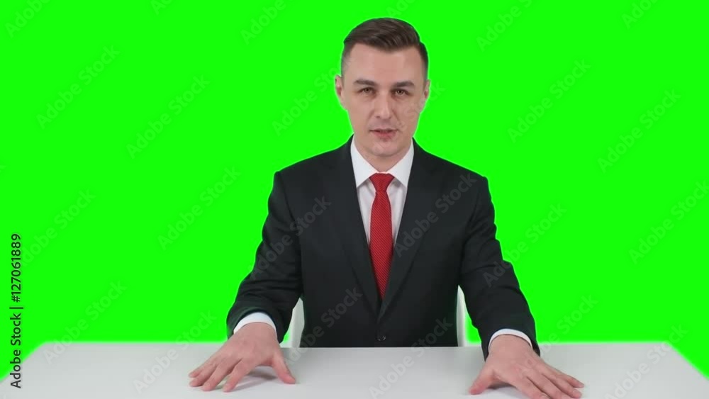 Man in suit sitting behind table gesticulating and showing something with his hands, green screen backdrop in background Man in suit sitting behind table gesticulating and showing something with his hands, green screen backdrop in background