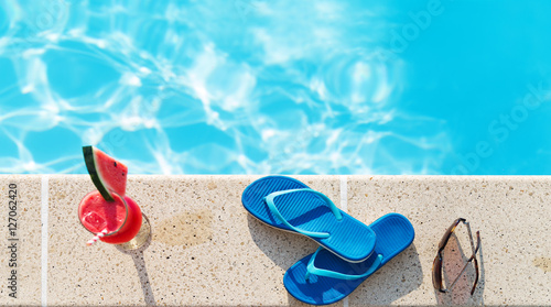 Glass of orange juice drink fresh with flowers, sunglasses and slippers on border of a swimming pool - holiday tropical concept