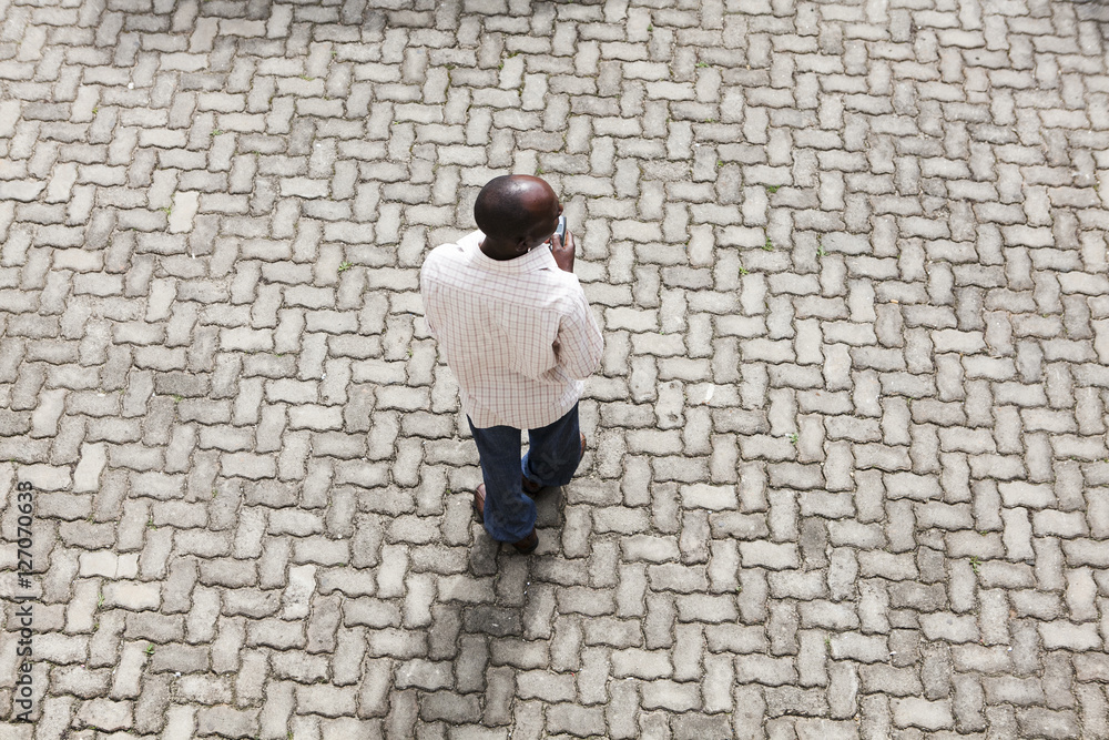 A Rwandan man seen from above holding a mobile phone whilst standing on a street made of bricks in a pattern. A Rwandan man seen from above holding a mobile phone whilst standing on a street made of bricks in a pattern.