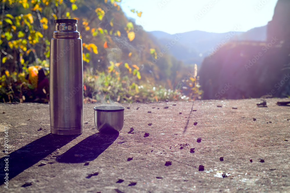 Thermos and cup with hot tea in sunny day with mountain background