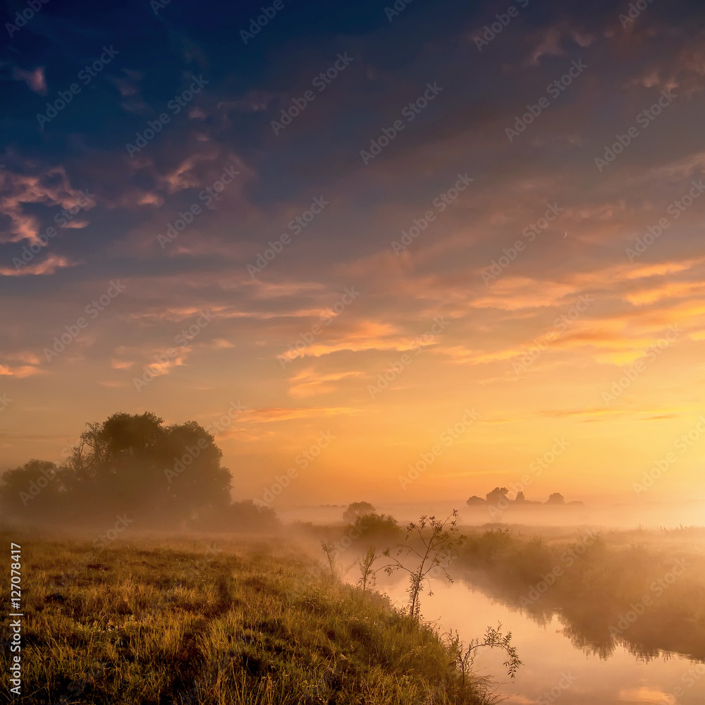 Fantastic foggy river with fresh grass in the sunlight. majestic misty sunrise with colorful clouds on the sky,    Dramatic unusual scene. Warm sundown over meadow. Beauty in the world.