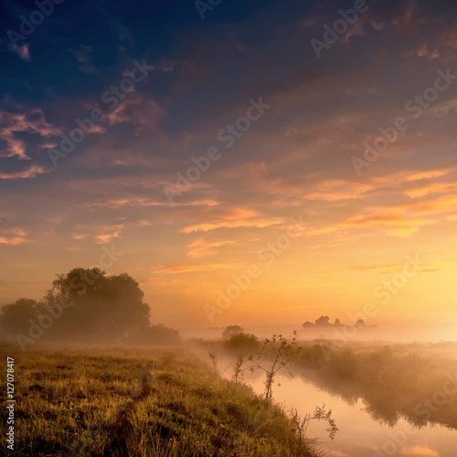 Fantastic foggy river with fresh grass in the sunlight. majestic misty sunrise with colorful clouds on the sky, Dramatic unusual scene. Warm sundown over meadow. Beauty in the world.