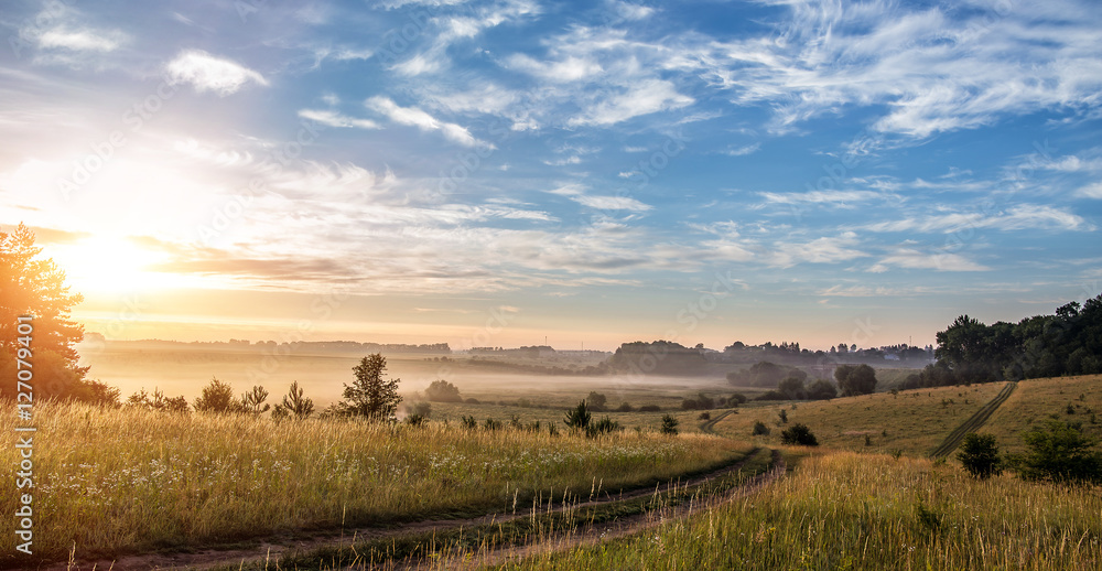 Fantastic foggy sunny day. ground road in the rural field with fresh grass in the sunlight. majestic misty sunrise with colorful clouds on the sky, Warm sundown over meadow. Fantastic foggy sunny day. ground road in the rural field with fresh grass in the sunlight. majestic misty sunrise with colorful clouds on the sky, Warm sundown over meadow.