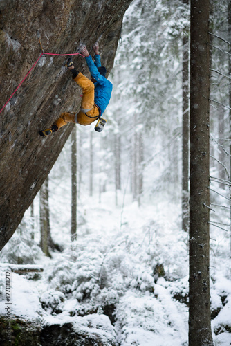 Extreme winter sport climbing. Young male rock climber on a rock wall. Snowy forest on the background,