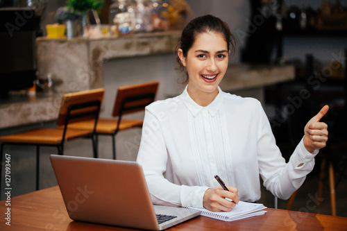 Young Caucasian woman showing thumbs up with laptop and notebook in the modern interior office - Gesture and thumbs up sign