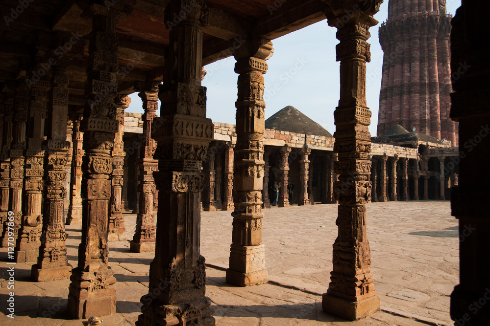 Ornate stone work on pillars lit by sun with next to plaza and Qutub Minar tower in background