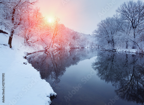 Winter forest on the river at sunset. Colorful landscape with snowy trees, beautiful frozen river with reflection in water, sun. Seasonal. Winter trees, lake and blue sky. Frosty snowy river. Weather