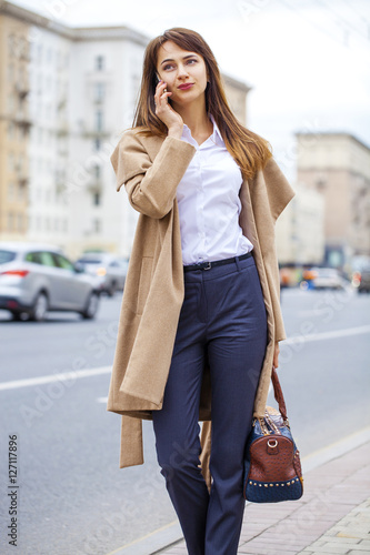 Portrait of happy young brunette woman in beige coat talking on © Andrey_Arkusha