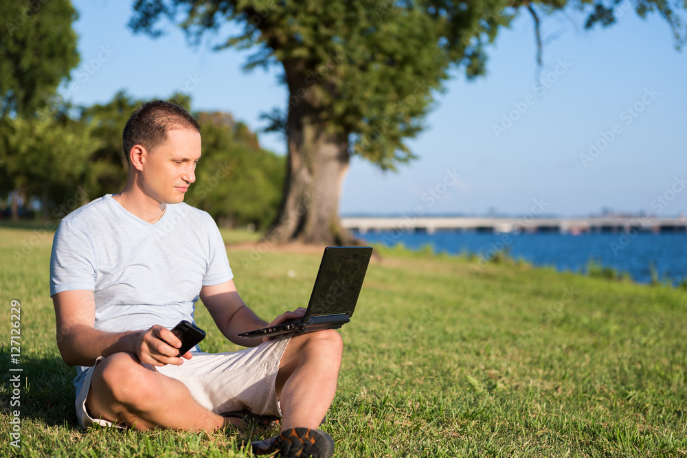 Young man looking at laptop and holding phone, sitting outside in green grass park Young man looking at laptop and holding phone, sitting outside in green grass park