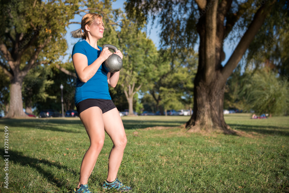 Young fit woman doing squats with heavy kettlebell in green outdoor park Young fit woman doing squats with heavy kettlebell in green outdoor park