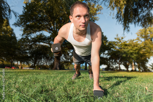 Young man performing dumbbell row exercise lifting 12 pound weight in green grass outdoor park