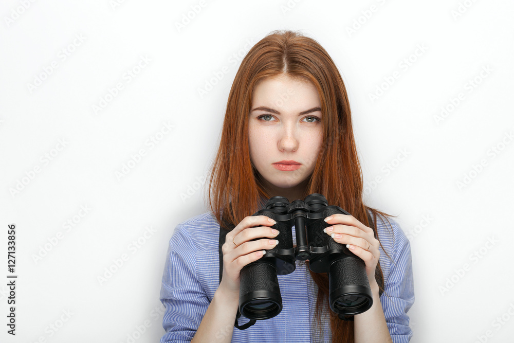 Portrait of cute redhead girl wearing blue striped shirt smiling with happiness and joy while posing with binoculars against white studio background