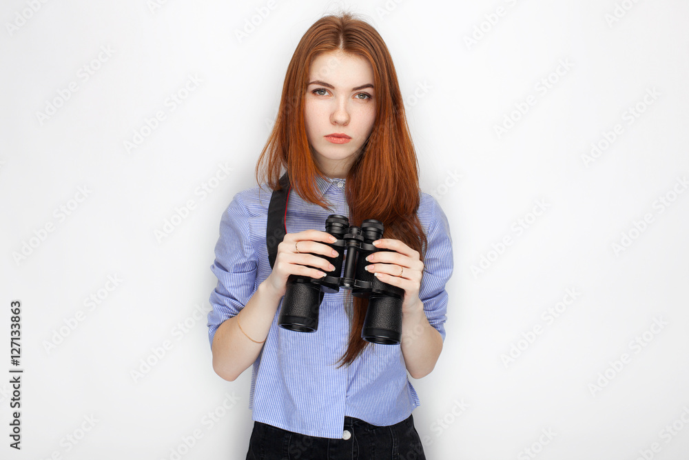 Portrait of cute redhead girl wearing blue striped shirt smiling with happiness and joy while posing with binoculars against white studio background Portrait of cute redhead girl wearing blue striped shirt smiling with happiness and joy while posing with binoculars against white studio background