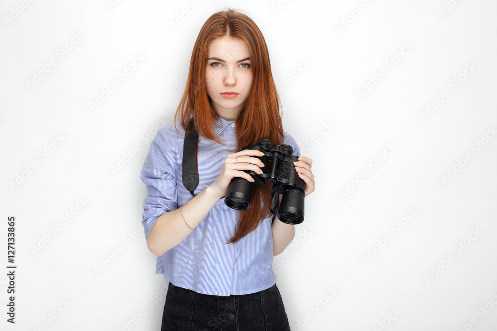 Portrait of cute redhead girl wearing blue striped shirt smiling with happiness and joy while posing with binoculars against white studio background